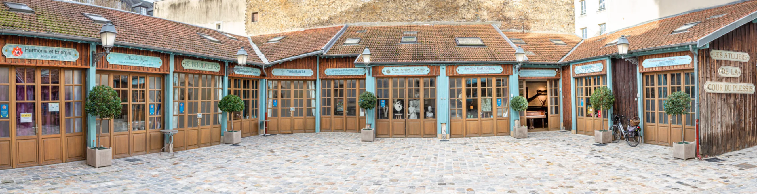 Cour pavée des Ateliers de la Cour Duplessis : façades en bois bleu clair, oliviers en pots