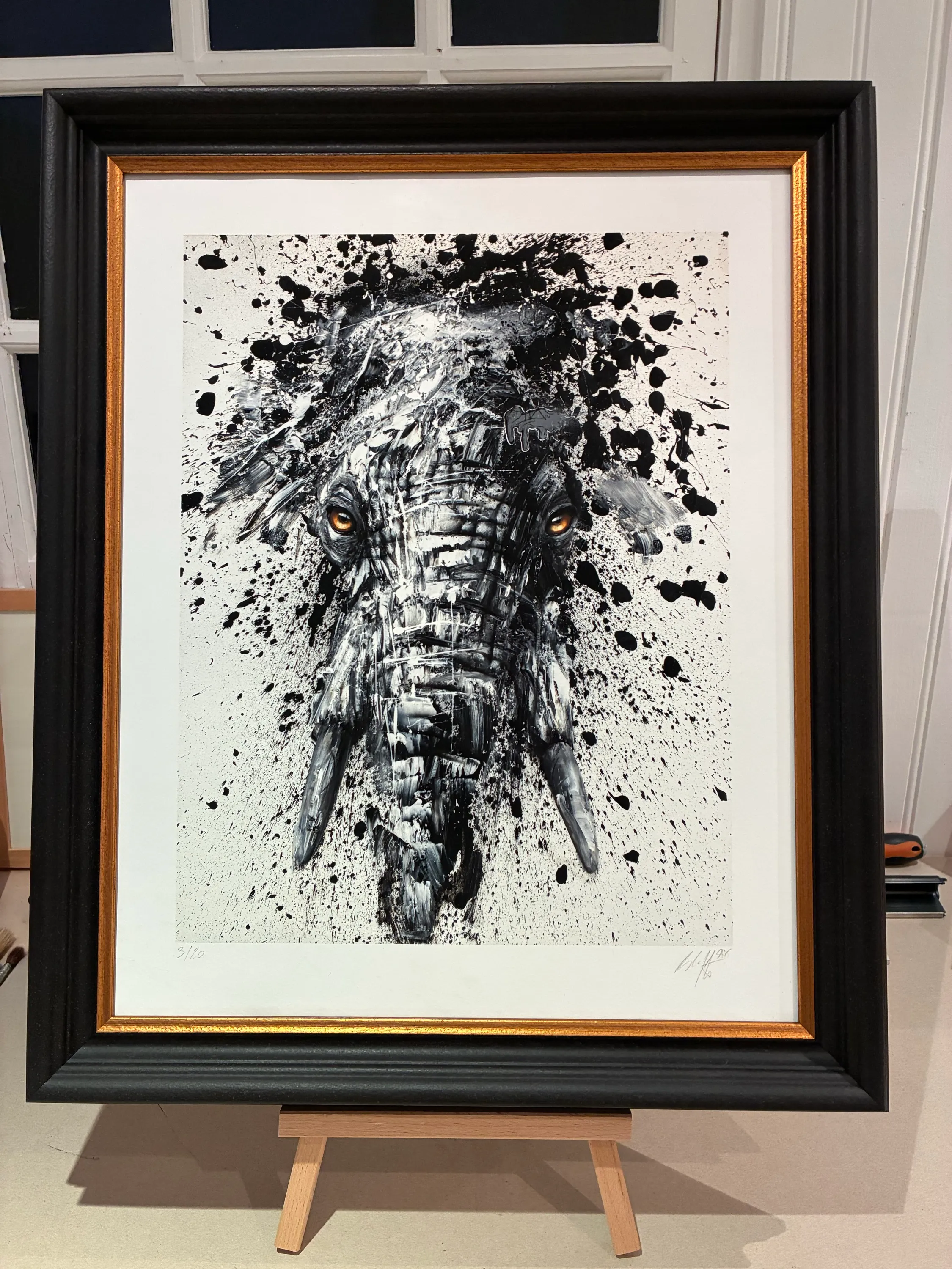Peinture à l'encre noire projetée sur papier blanc, faisant apparaître le visage d'un éléphant aux yeux dorés, encadrée dans un cadre noir laqué bordé d'un fin filet doré intérieur
