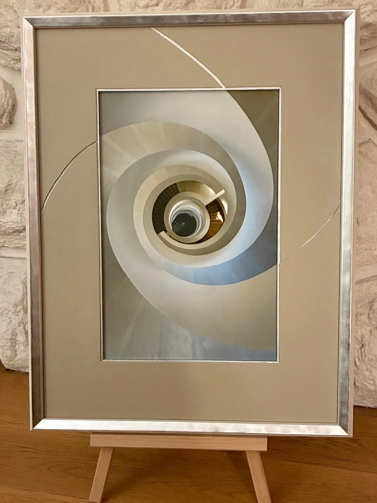 Photographie d'un escalier en spirale vu d'en bas, dans un cadre argenté avec passe-partout aux découpes courbes